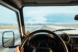 A driver’s perspective from inside a moving vehicle, showing a vast road stretching into the distance bordered by snow-covered plains and a mountain range under a partly cloudy sky. The dashboard and steering wheel are visible, indicating a focus on travel or journey.
