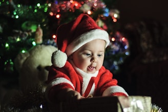 A baby dressed in a Santa Claus outfit is reaching out towards a gift, with a Christmas tree decorated with colorful lights in the background. A large white teddy bear is partially visible behind the child.