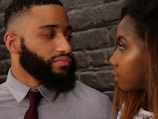 A man and a woman are looking at each other closely in front of a textured brick wall. The man has a full beard and is wearing a grey shirt with a red tie. The woman has long hair and is dressed in a white top. Their expressions are serious, suggesting an intense moment.