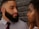 A man and a woman are looking at each other closely in front of a textured brick wall. The man has a full beard and is wearing a grey shirt with a red tie. The woman has long hair and is dressed in a white top. Their expressions are serious, suggesting an intense moment.