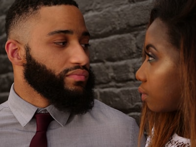 A man and a woman are looking at each other closely in front of a textured brick wall. The man has a full beard and is wearing a grey shirt with a red tie. The woman has long hair and is dressed in a white top. Their expressions are serious, suggesting an intense moment.