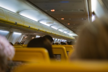 The interior of an airplane cabin features a row of overhead luggage compartments with visible exit signs. Passengers are seated, with bright yellow headrests prominently displayed. The ceiling lights are softly illuminated, creating a calm ambiance within the cabin.