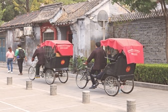 Two rickshaw drivers are cycling on a paved street lined with bollards. The rickshaws have red canopies and are numbered 20 and 16. Pedestrians walk nearby alongside old, weathered buildings with traditional tiled roofs.