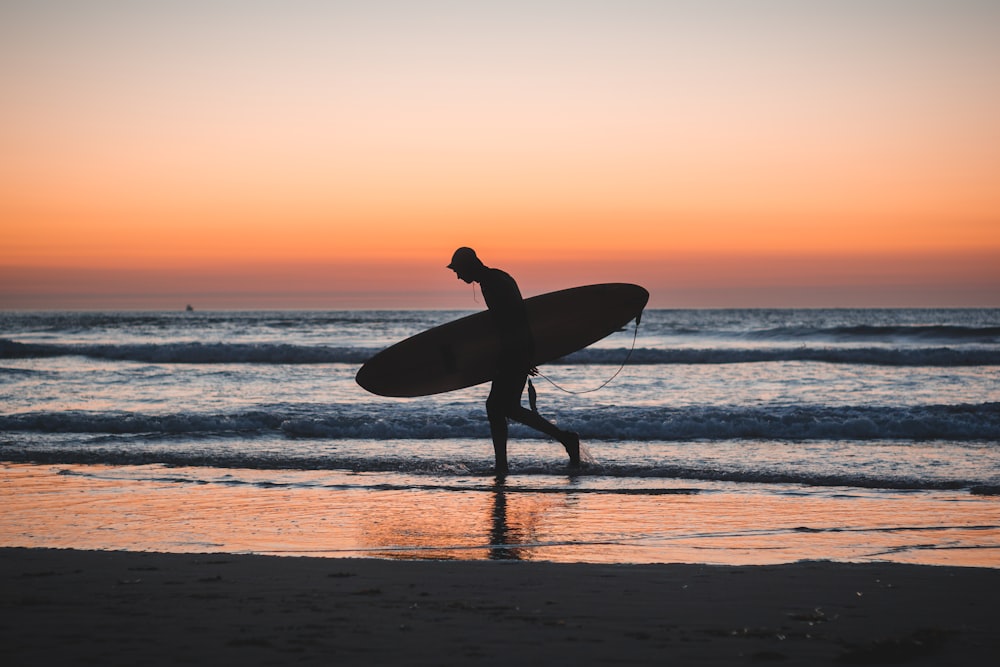 silhouette photo of person holding surfboard near shore