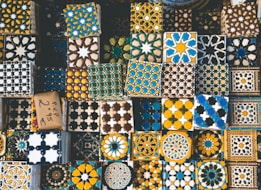 A collection of decorative tiles featuring intricate geometric and floral patterns in a variety of colors such as blue, yellow, black, and white. The tiles are arranged closely together, showcasing a rich tapestry of cultural designs and artistic craftsmanship.