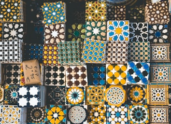 A collection of decorative tiles featuring intricate geometric and floral patterns in a variety of colors such as blue, yellow, black, and white. The tiles are arranged closely together, showcasing a rich tapestry of cultural designs and artistic craftsmanship.