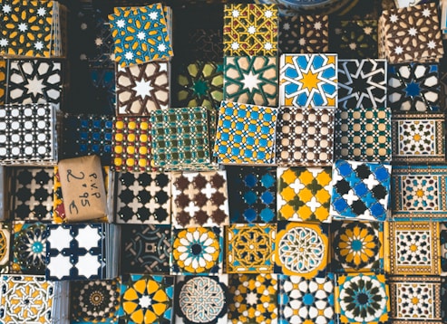 A collection of decorative tiles featuring intricate geometric and floral patterns in a variety of colors such as blue, yellow, black, and white. The tiles are arranged closely together, showcasing a rich tapestry of cultural designs and artistic craftsmanship.