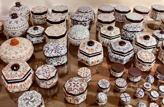 A collection of intricately patterned ceramic containers displayed on a flat surface. Each container features geometric and floral designs in various colors, primarily focused on shades of blue, orange, and green. The containers vary in size and shape, with hexagonal and cylindrical pieces, all topped with a dark-colored knob.