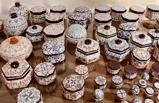 A collection of intricately patterned ceramic containers displayed on a flat surface. Each container features geometric and floral designs in various colors, primarily focused on shades of blue, orange, and green. The containers vary in size and shape, with hexagonal and cylindrical pieces, all topped with a dark-colored knob.