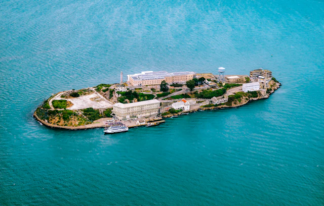 Aerial view of Alcatraz Island showing the old federal prison buildings surrounded by turquoise bay water.