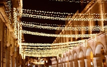 String lights are beautifully strung across a street between buildings, creating an enchanting canopy. The architecture features arches and columns, illuminated softly by the warm glow of the lights. The scene appears to be in a historic or old town setting, with a festive and welcoming atmosphere.