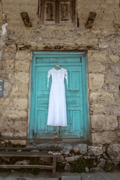 A white lace wedding dress hangs on a turquoise wooden door set against a rustic stone wall. The scene includes an old, weathered bench and a small bouquet of flowers lying on it.