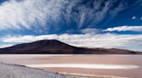 A vast landscape with a large salt flat and a distant mountain range under a dramatic cloudy sky. The salt flat is reflecting hues of pink and white, creating a serene atmosphere.