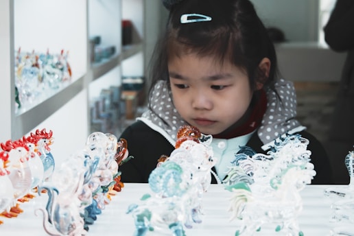 A young girl intently examines a collection of small, colorful, glass figurines displayed on a shelf. Her expression is one of focus and curiosity. The figurines are intricately detailed with a variety of colors and designs.