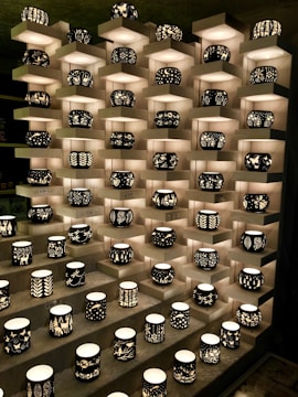 A display of intricately designed candleholders arranged on tiered shelves. Each holder features unique black silhouettes of various patterns, including nature scenes and abstract designs, illuminated from within by soft light.