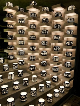 A display of intricately designed candleholders arranged on tiered shelves. Each holder features unique black silhouettes of various patterns, including nature scenes and abstract designs, illuminated from within by soft light.