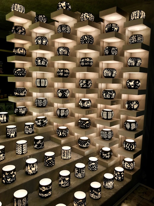 A display of intricately designed candleholders arranged on tiered shelves. Each holder features unique black silhouettes of various patterns, including nature scenes and abstract designs, illuminated from within by soft light.