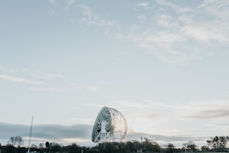 A large radio telescope dish is situated in a landscape with trees along the horizon. The sky above is vast and partly cloudy, creating a serene and open atmosphere.