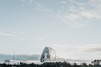 A large radio telescope dish is situated in a landscape with trees along the horizon. The sky above is vast and partly cloudy, creating a serene and open atmosphere.