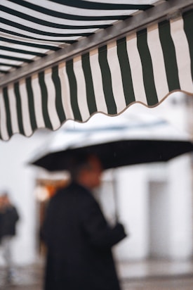 A striped awning appears prominently in the foreground, consisting of alternating black and white stripes. In the background, a person holds an umbrella, with the figure blurred and out of focus, suggesting a rainy day atmosphere.