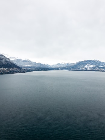 An expansive drone panorama of a serene lake reflecting the snow-capped Rockies under a bright sky.