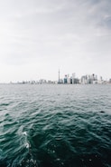 A skyline featuring modern high-rise buildings with a prominent tower in the center, viewed across a large body of water under a cloudy sky.