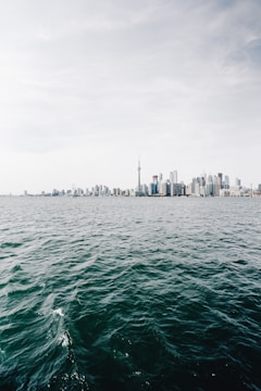 A skyline featuring modern high-rise buildings with a prominent tower in the center, viewed across a large body of water under a cloudy sky.