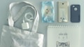 A neatly arranged collection of items on a flat surface, including a canvas tote bag, a white power adapter and cable, a package with a cute animal face design, two smartphones, one of which has a gold color and a transparent back, a black phone case, and a spiral-bound notebook with a cat-themed cover.