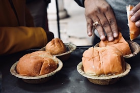 A close-up view of several bread rolls placed in biodegradable paper containers. One person is interacting with the rolls, their hand visible and wearing a ring. The focus is on the food, with the hand and background slightly blurred.