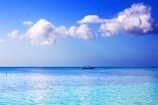 A tranquil seascape with calm turquoise waters under a vibrant blue sky. Fluffy white clouds float above, and a lone boat is gently positioned at the horizon.