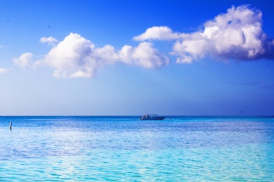 A tranquil seascape with calm turquoise waters under a vibrant blue sky. Fluffy white clouds float above, and a lone boat is gently positioned at the horizon.