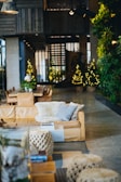 A cozy and stylish living room with a tan leather sofa adorned with soft, fluffy cushions. There is a large wooden table with chairs in the background, accompanied by warm yellow fairy lights shaped like Christmas trees. The walls are decorated with lush green plants, adding a natural element to the space. Modern lighting fixtures hang from the ceiling, providing a warm and inviting ambiance.