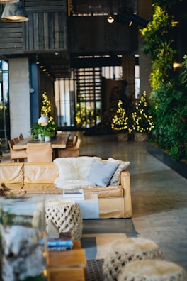A cozy and stylish living room with a tan leather sofa adorned with soft, fluffy cushions. There is a large wooden table with chairs in the background, accompanied by warm yellow fairy lights shaped like Christmas trees. The walls are decorated with lush green plants, adding a natural element to the space. Modern lighting fixtures hang from the ceiling, providing a warm and inviting ambiance.