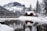 A snowy landscape with a wooden cabin surrounded by tall evergreen trees, situated near a tranquil body of water. Snow-covered mountains form a majestic backdrop, reflecting on the calm surface of the lake.