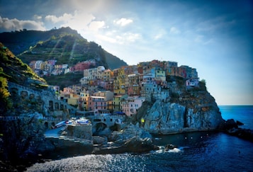 A picturesque coastal village with colorful buildings perched atop a rugged cliff overlooking the sea. The sun casts rays through a partly cloudy sky, illuminating the vibrant architecture and lush greenery on the hillside.