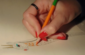 A hand with red painted nails is holding an orange and green colored pencil, coloring a detailed drawing of an animal's head with intricate patterns. The image shows the person filling in orange on a section of the design while the rest of the drawing remains uncolored.