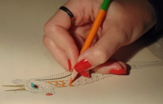 A hand with red painted nails is holding an orange and green colored pencil, coloring a detailed drawing of an animal's head with intricate patterns. The image shows the person filling in orange on a section of the design while the rest of the drawing remains uncolored.
