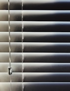 Horizontal blinds partially closed with minimal light seeping through, creating a pattern of alternating light and shadow. A pull cord hangs on the left side of the blinds.