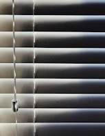 Horizontal blinds partially closed with minimal light seeping through, creating a pattern of alternating light and shadow. A pull cord hangs on the left side of the blinds.