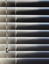 Horizontal blinds partially closed with minimal light seeping through, creating a pattern of alternating light and shadow. A pull cord hangs on the left side of the blinds.