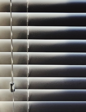 Horizontal blinds partially closed with minimal light seeping through, creating a pattern of alternating light and shadow. A pull cord hangs on the left side of the blinds.