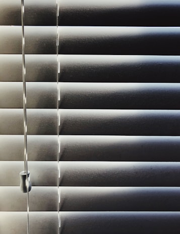 Horizontal blinds partially closed with minimal light seeping through, creating a pattern of alternating light and shadow. A pull cord hangs on the left side of the blinds.