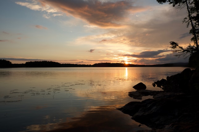A serene lakeside view at sunset, with calm waters reflecting the warm hues of the sky. The horizon is dotted with distant tree silhouettes, while patches of clouds catch the golden and orange shades of the setting sun. In the foreground, a rocky shore with sparse vegetation frames the scene, adding depth and contrast.