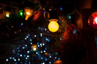 Various colorful light bulbs and decorations are visible, with a prominent glowing yellow bulb in the foreground surrounded by green foliage and red tinsel. In the background, blue and white lights form a bokeh effect, adding a festive atmosphere.