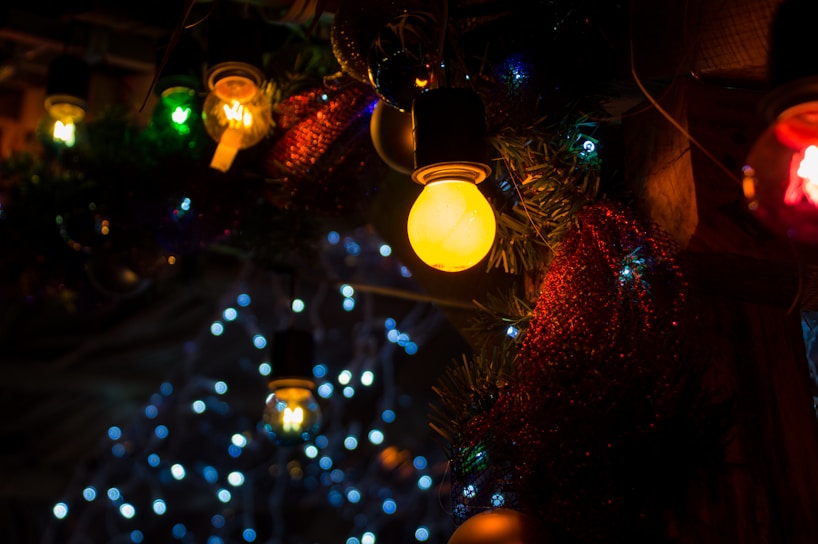 Various colorful light bulbs and decorations are visible, with a prominent glowing yellow bulb in the foreground surrounded by green foliage and red tinsel. In the background, blue and white lights form a bokeh effect, adding a festive atmosphere.
