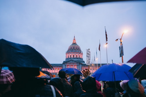 A crowd of diverse citizens holding umbrellas, standing firm against a rain of money.