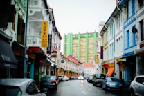 A vibrant city street lined with colorful buildings and various business signs. The street is wet, likely from recent rain, with several parked cars on either side. In the background, a modern residential or commercial high-rise building is visible with a distinct green color.
