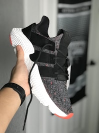 A hand holds a stylish sneaker with a black and white knit upper, featuring black straps and red accents. The shoe has a distinctive textured white midsole. There's a blurred background of a door and wall with unclear details.