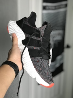 A hand holds a stylish sneaker with a black and white knit upper, featuring black straps and red accents. The shoe has a distinctive textured white midsole. There's a blurred background of a door and wall with unclear details.