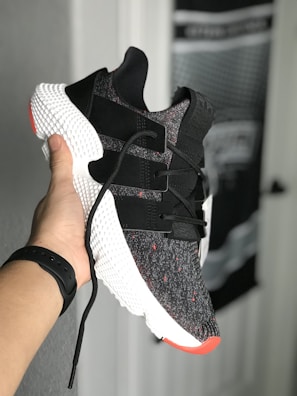 A hand holds a stylish sneaker with a black and white knit upper, featuring black straps and red accents. The shoe has a distinctive textured white midsole. There's a blurred background of a door and wall with unclear details.